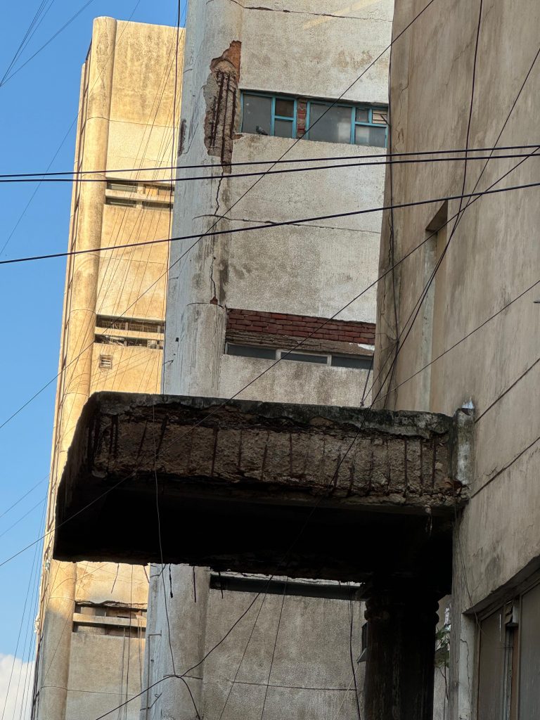 Wide shot showing cracked façade, dangling cables and deteriorated beam above service lane.